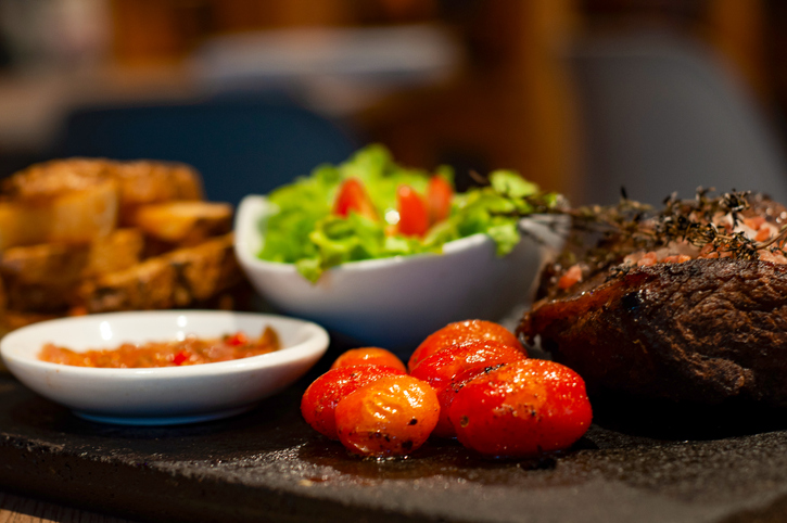 A perfectly cooked steak sits on a black stone plate, exuding a savory aroma, accompanied by a plate of fresh green salad studded with red cherry tomatoes and a side of roasted golden potatoes.